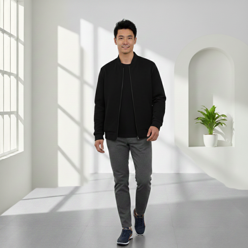 Man in black jacket and gray pants standing in a bright, minimalistic room with a plant.
