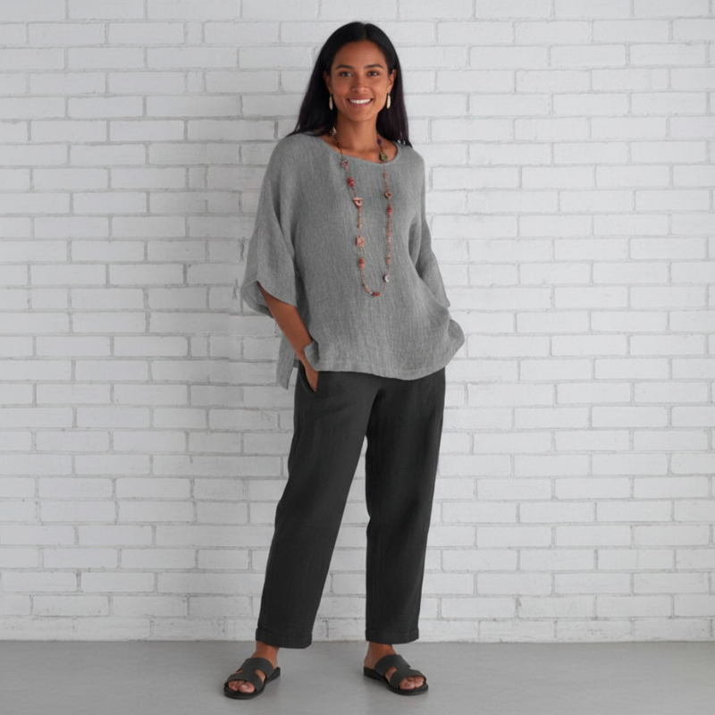 Woman wearing a gray sweater and dark pants standing against a white brick wall.