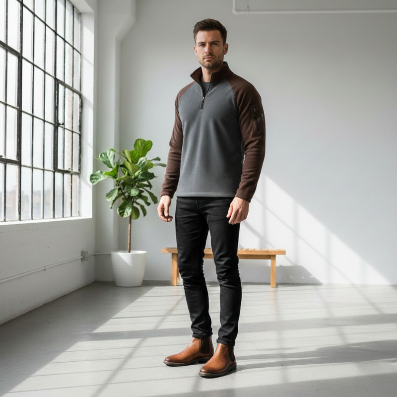 Man wearing a brown and gray pullover in a bright room with large windows and a plant.