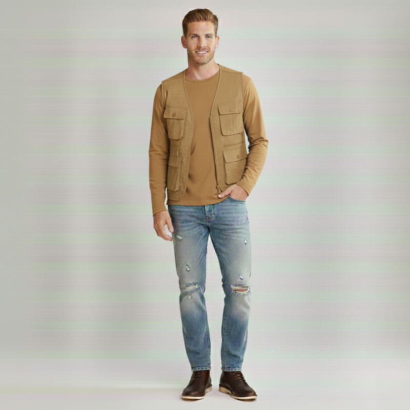 Man wearing a casual vest layered over a tan sweater and jeans in a clean studio environment.