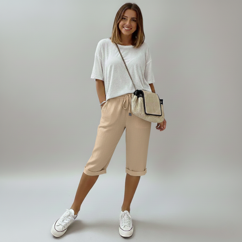 Woman wearing a white t-shirt, beige shorts, and white sneakers, holding a straw bag against a plain background.