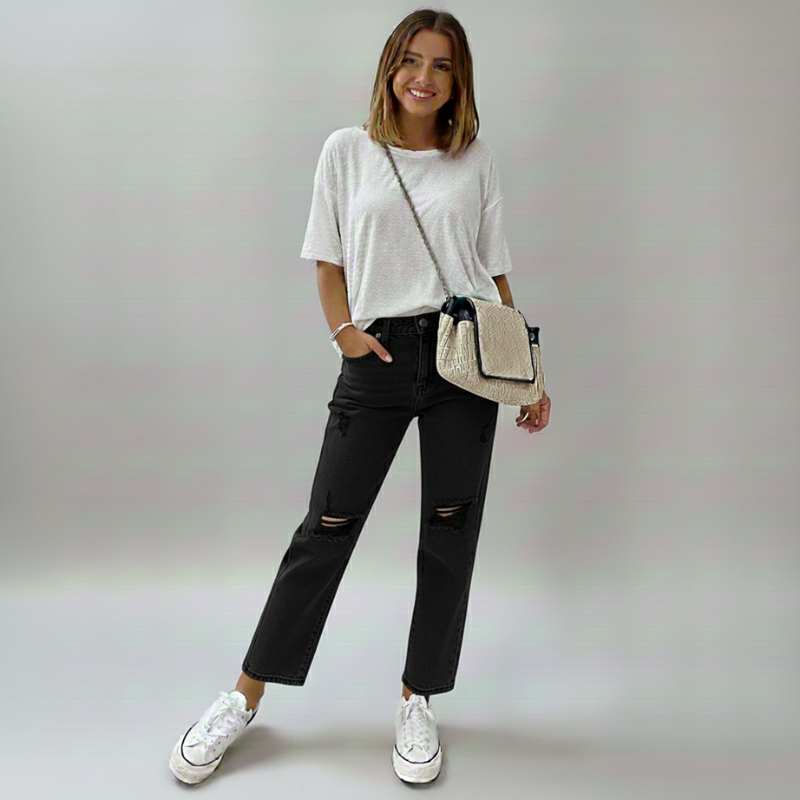 Woman wearing a white oversized shirt, black ripped jeans, and white sneakers, holding a beige handbag on a plain background.