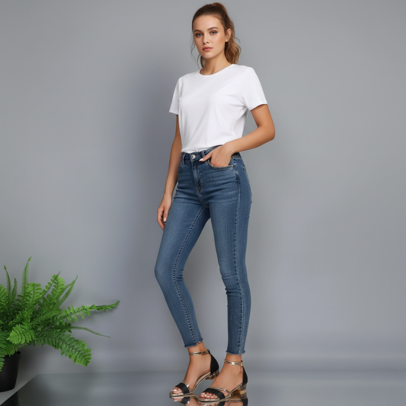 Woman wearing blue jeans and a white t-shirt on a gray background with a plant to the left.