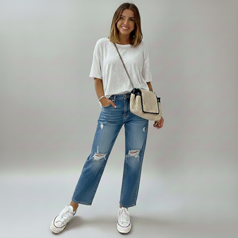 Woman wearing a white t-shirt, blue jeans, and white sneakers, holding a beige crossbody bag on a plain background.