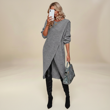 Woman wearing a gray sweater dress with black leggings and boots, holding a phone and a handbag on a beige background.