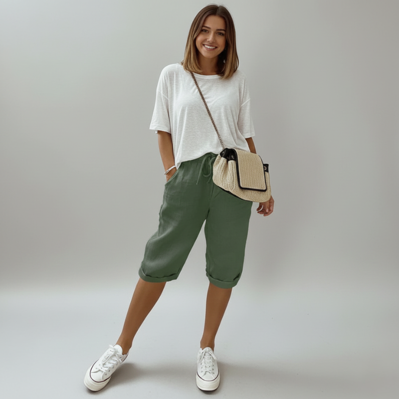 Woman wearing a white top, green pants, and white sneakers, holding a beige handbag on a plain background