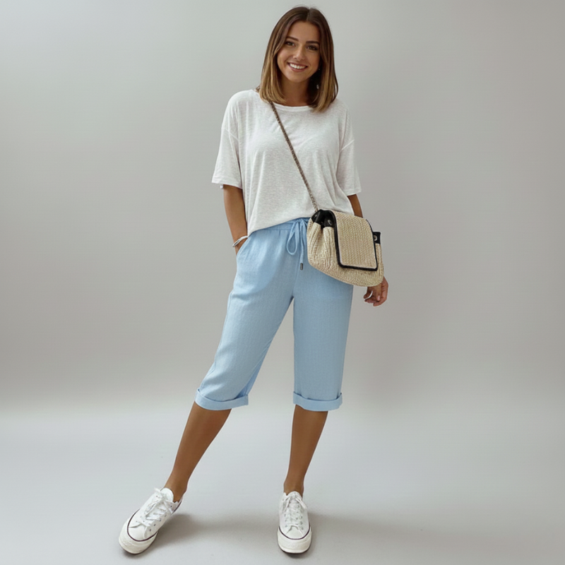 Woman wearing a white t-shirt, light blue jeans, and white sneakers, holding a beige crossbody bag against a plain background.