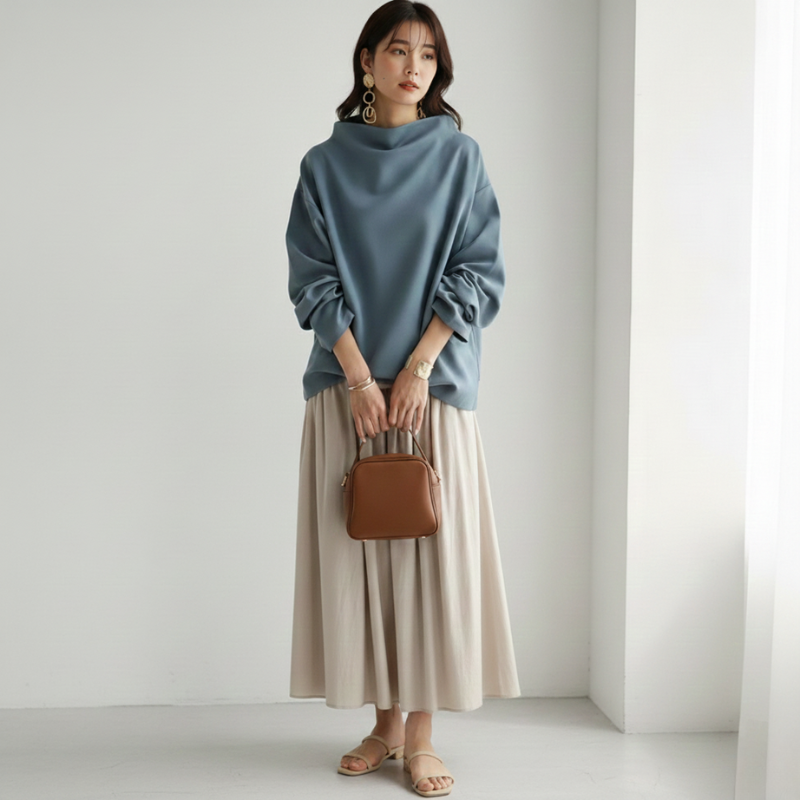 Woman wearing a blue sweater and beige skirt holding a brown handbag against a white background