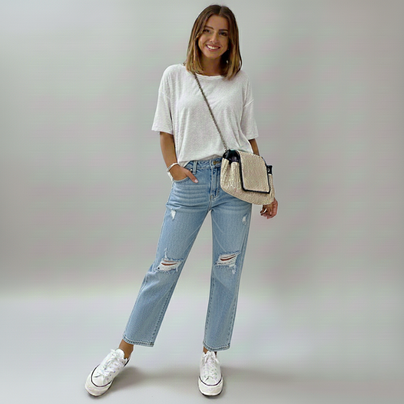 Woman wearing a white t-shirt, blue jeans, and white sneakers, holding a beige crossbody bag on a plain background.